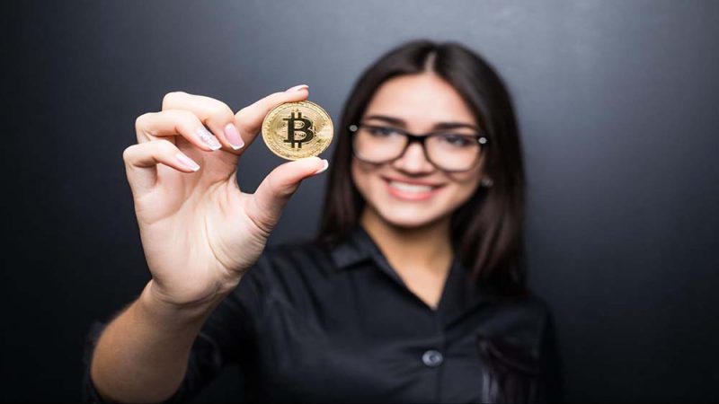 A businesswoman dressed in black holding a Bitcoin coin with her thumb and index finger.